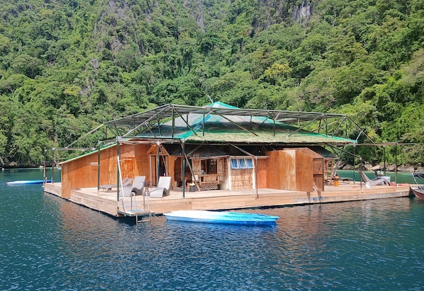 Une structure flottante en bois, dotée d’une terrasse couverte et de chaises longues, entourée d’une étendue d’eau calme et d’une forêt dense.