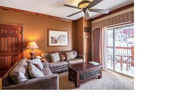 A living room with a dark wooden coffee table, a brown leather sofa, a wooden door, and a ceiling fan. - Two bedroom Two Bath with private lock-off unit (Park City)