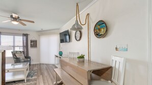 Un coin repas avec une table en bois, des bancs et des chaises. On y trouve également un ventilateur de plafond, un téléviseur fixé au mur et une horloge.
