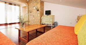 A room with a wooden table, a sofa, a television on a dresser, and a stone wall.