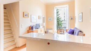 Un salon moderne doté d’un îlot en marbre, d’un téléviseur à écran plat et d’un coin repas avec une table et des chaises. Un escalier est visible sur la gauche et mène à un autre niveau.