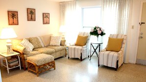 Ein Wohnzimmer mit einer Couch, zwei Sesseln, einem kleinen Tisch und einer Lampe. An der Wand hängen drei gerahmte Bilder.