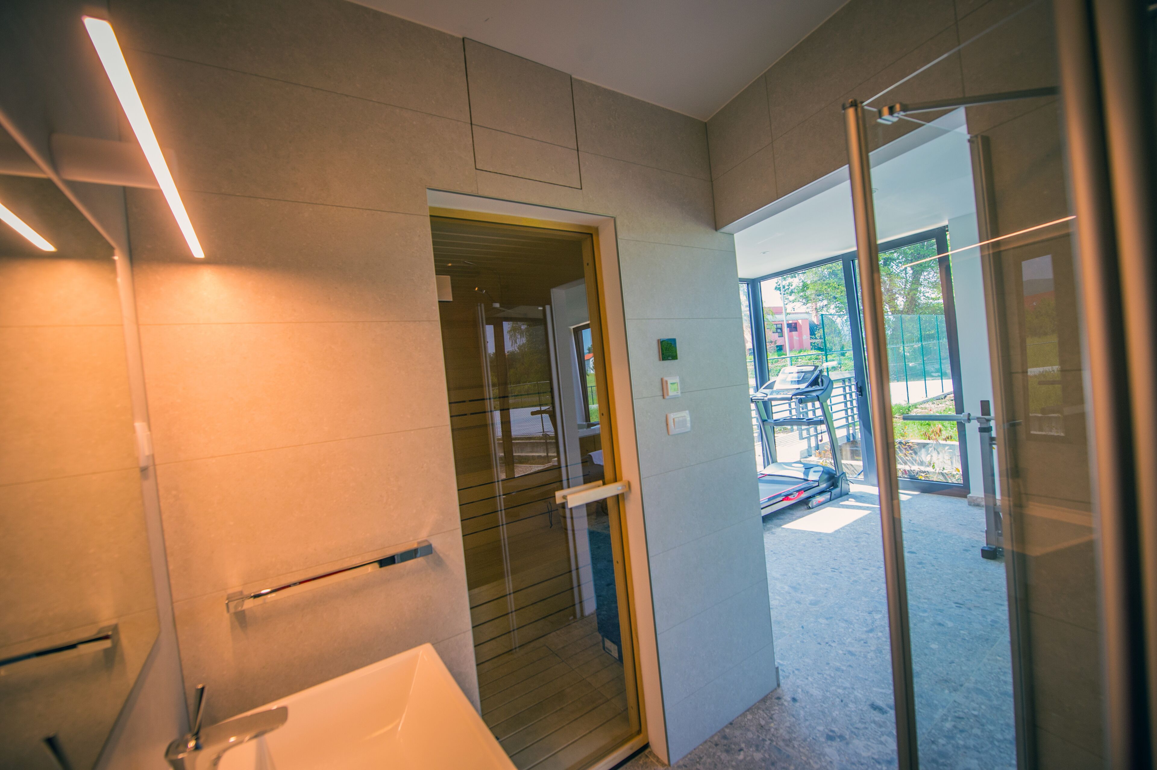 A modern bathroom with a glass shower enclosure, a white sink, and a door leading to an outdoor area with a treadmill.