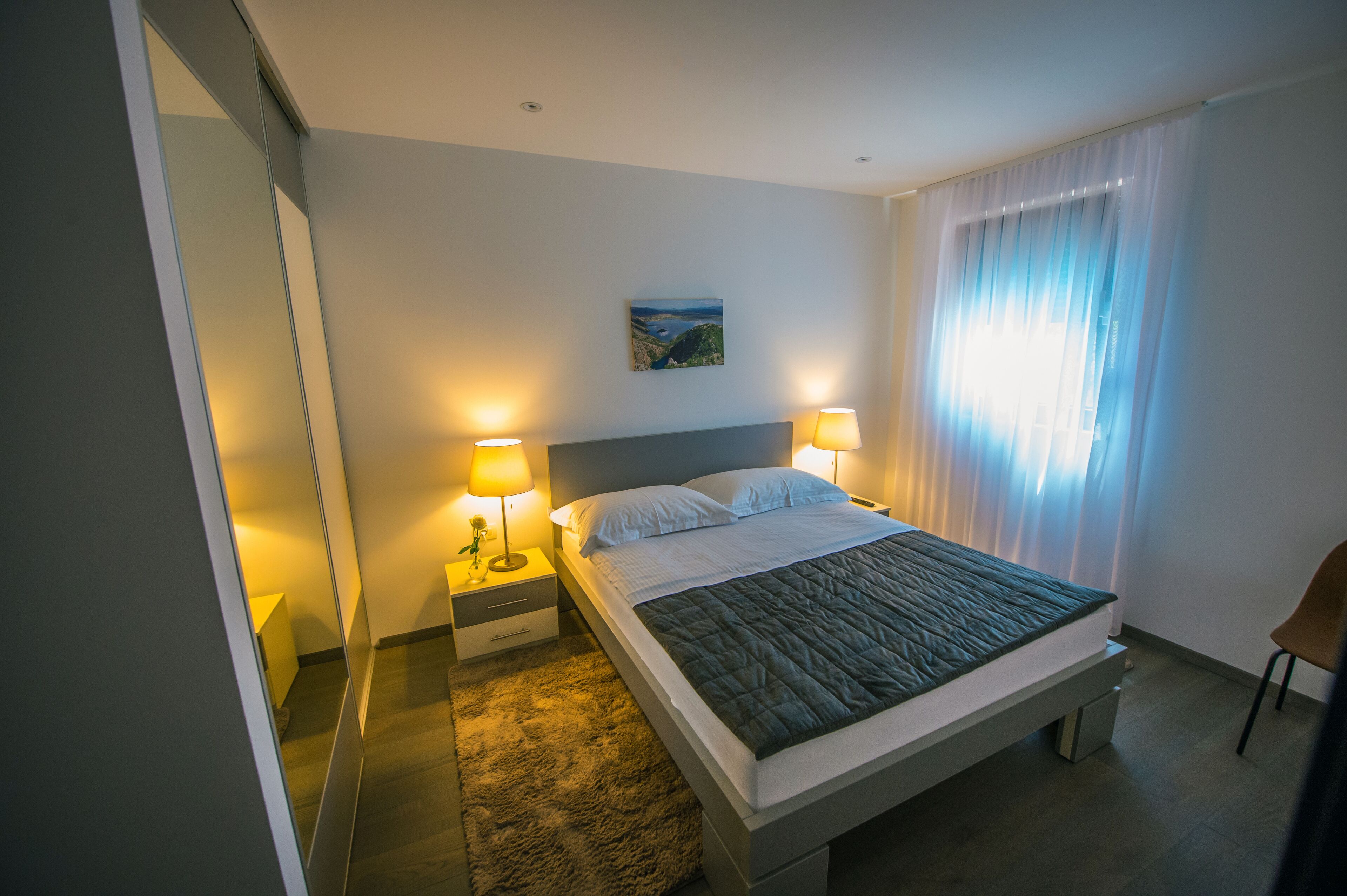 Quarto de hotel moderno com uma cama grande, abajures, um quadro na parede e vista para o exterior através de cortinas leves.
