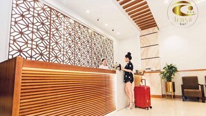 A hotel reception area with a modern design, featuring a geometric patterned wall, a wooden reception desk, and a red suitcase on the floor.