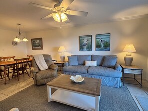 A living room with a sofa, armchairs, a coffee table, a dining table with chairs, a ceiling fan, and wall-mounted lamps.