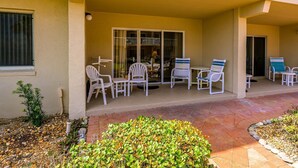 A covered outdoor patio with white plastic chairs and a table, a small garden area with shrubs, and a building entrance with a glass door.