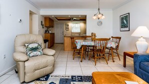 A living room with a beige sofa, a blue armchair, a wooden coffee table, and a dining area with a table and chairs.