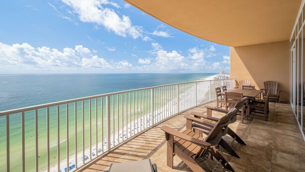 A balcony with a view of the ocean and beach, furnished with wooden chairs and tables.