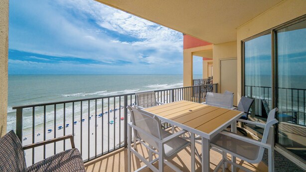 A balcony with a wooden table and white chairs, overlooking a beach with umbrellas and people.
