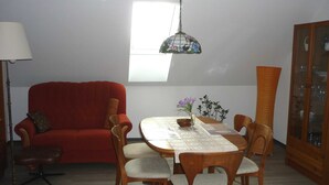 Un comedor con una mesa lista para cuatro personas, rodeada de sillas de madera. Un sofá rojo está colocado contra la pared y un jarrón alto y estrecho sobre la mesa.