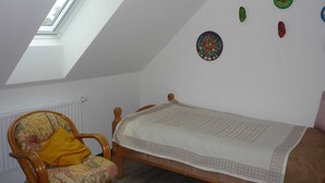 Una habitación pequeña y acogedora con techo inclinado, una cama individual, una silla de madera con cojín floral y un radiador.