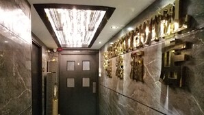 A modern elevator with a geometric light fixture above, a marble floor with a central design, and a wall with a metallic, abstract art piece.
