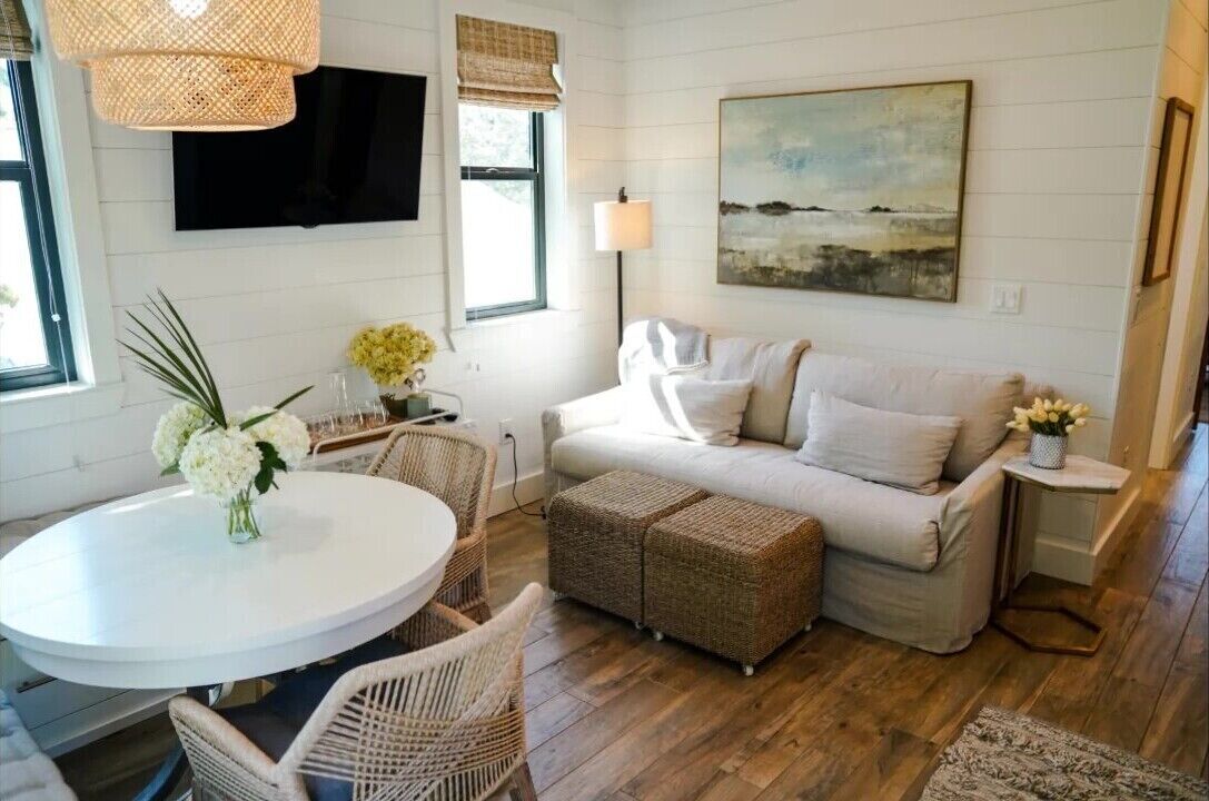 A cozy living room with a white sofa, wicker ottomans, a round dining table with chairs, a flat-screen TV, and a painting on the wall.