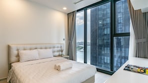 A modern hotel room with a large bed, a bedside table, and a view of a cityscape through floor-to-ceiling windows. - NES House - Riverview Apartment (Ho Chi Minh City)