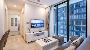 A modern living room with a flat-screen TV, a sofa, a coffee table, and a large window offering a city view. - NES House - Riverview Apartment (Ho Chi Minh City)
