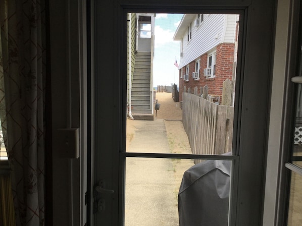 Visão através de uma porta para uma área externa estreita, com caminho de concreto, cerca de madeira e um prédio de tijolos com janela.