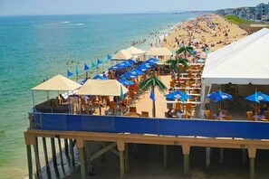 A beachside pier with outdoor seating and umbrellas, overlooking a crowded beach. - 🏖️Steps 2 the BEACH Cute &Cozy Retreat Oceanside w/ Marsh Views only w/ A/C &1 PRK☀️ (Salisbury)