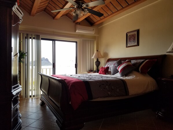 A bedroom with a bed, a ceiling fan, a window with a view, and a door.