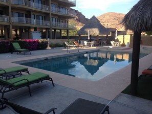A pool area with lounge chairs, a thatched-roof structure, and a building with balconies.