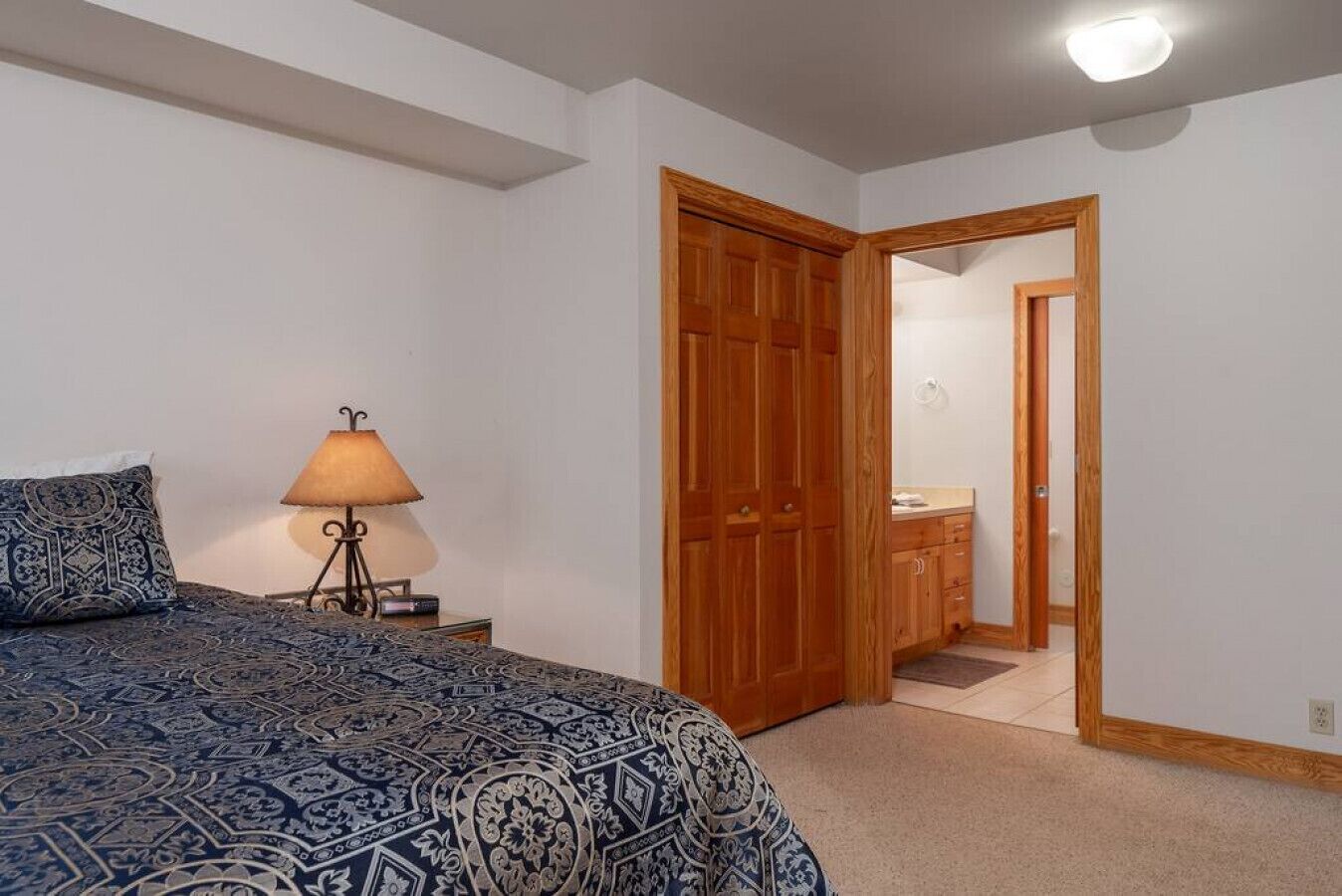 A bedroom with a bed, a nightstand with a lamp, a wooden wardrobe, and an open door leading to a bathroom.