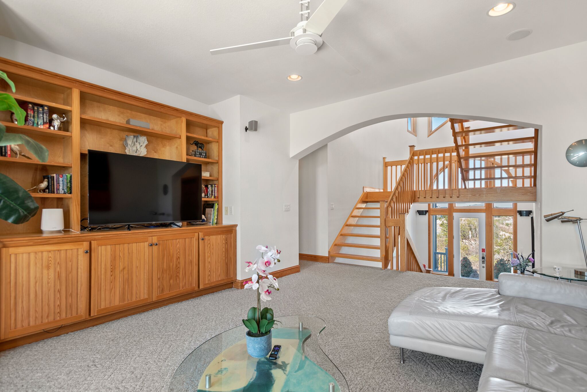 A modern living room with a flat-screen TV, wooden shelves, a glass coffee table with a plant, a white leather sofa, and a staircase leading to an upper level.
