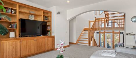 A modern living room with a flat-screen TV, wooden shelves, a glass coffee table with a plant, a white leather sofa, and a staircase leading to an upper level.