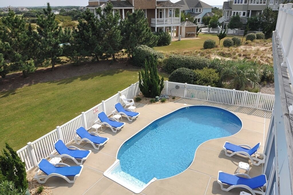 A swimming pool with lounge chairs, surrounded by a white fence and greenery.