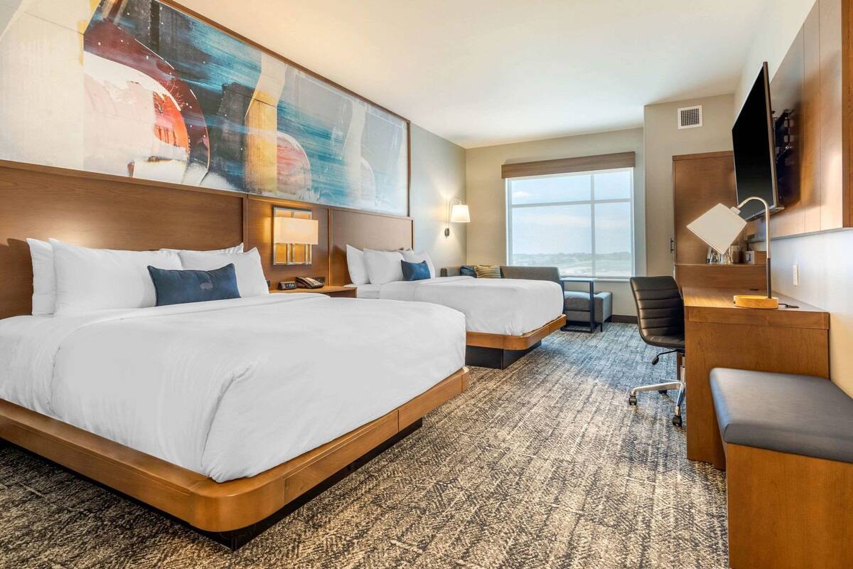 A hotel room with two beds, a desk, a chair, a TV, and a large window.