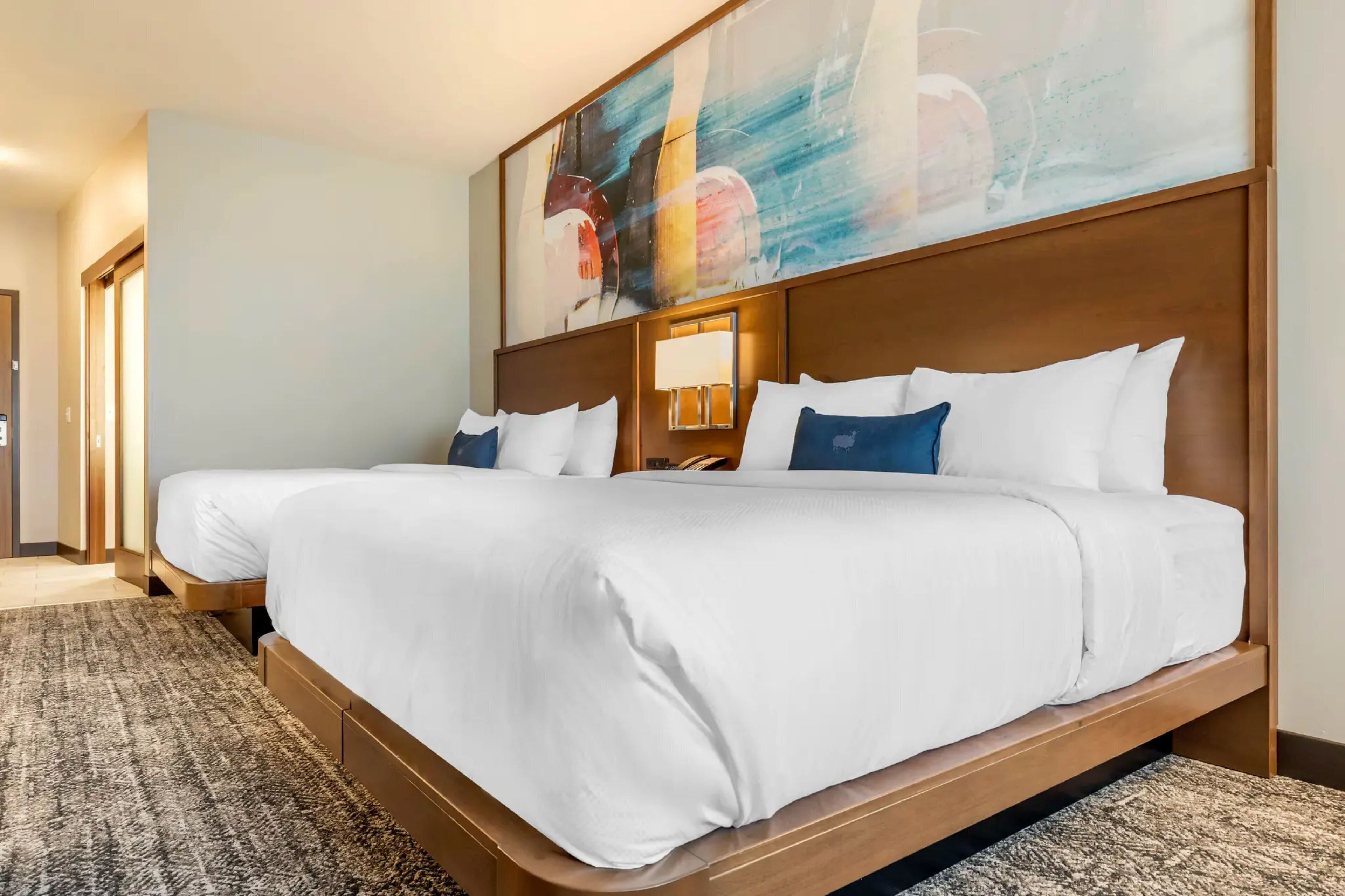 Ein Hotelzimmer mit zwei Betten, einem großen abstrakten Gemälde, einem Holzkopfteil und einem Teppichboden.