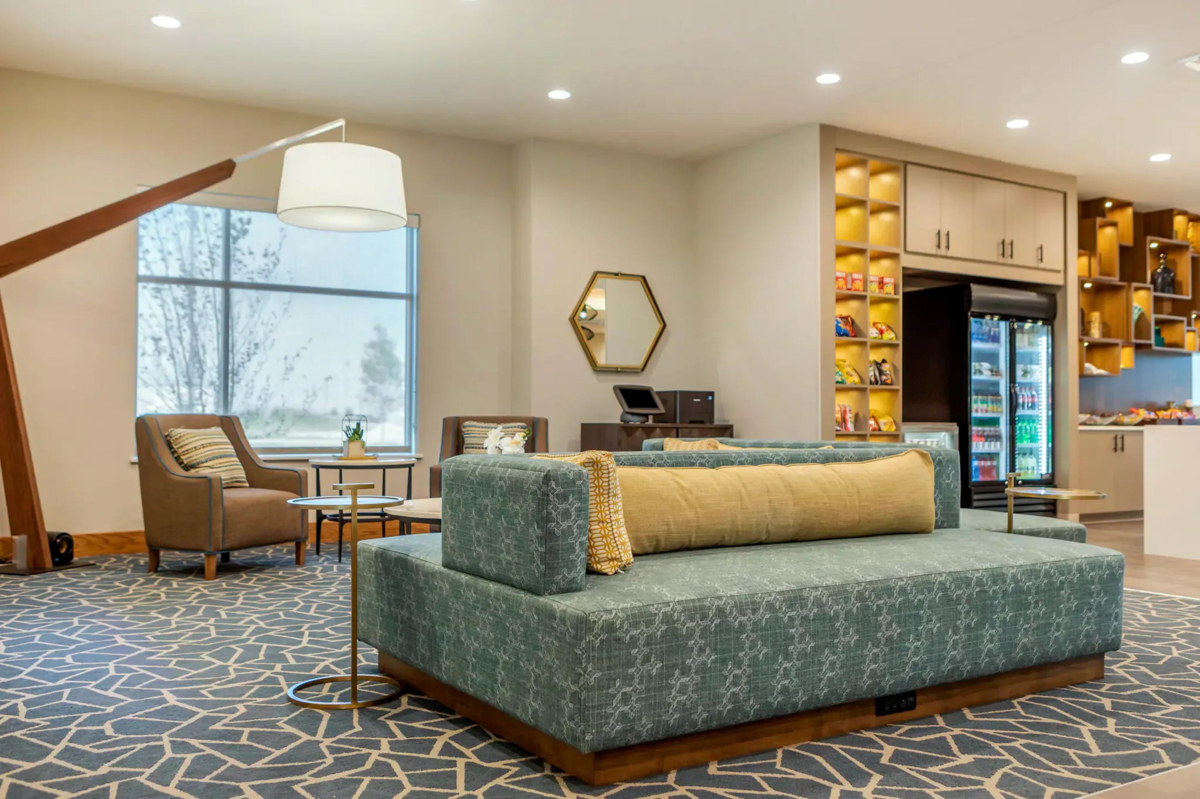 Habitación de hotel con sofá, sillón y una mesita con lámpara. Cuenta con un ventanal grande y un refrigerador con estantes.