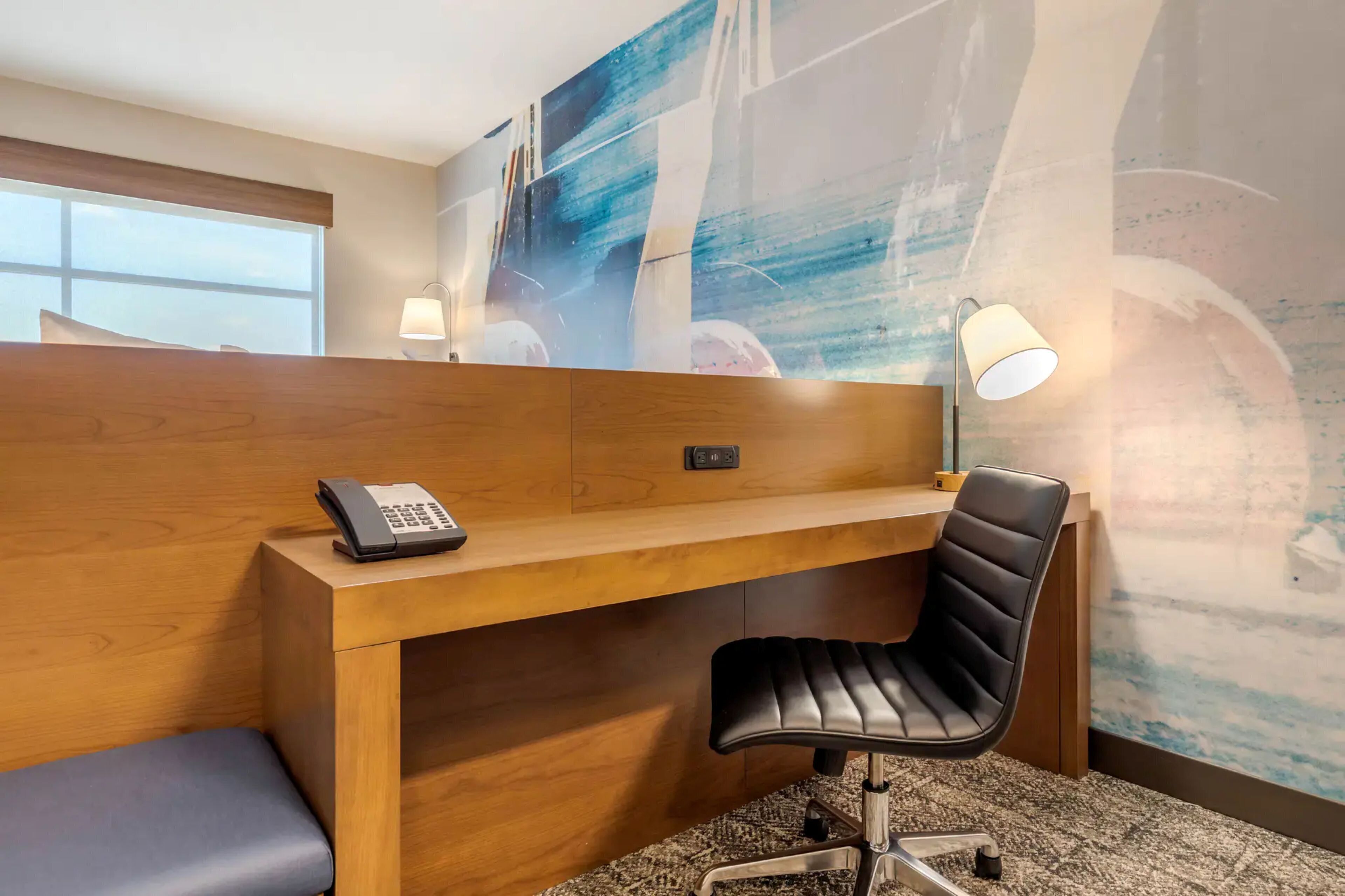 Une chambre d’hôtel avec un bureau en bois, un fauteuil de bureau noir, un téléphone et un mur orné d’œuvres d’art abstraites.