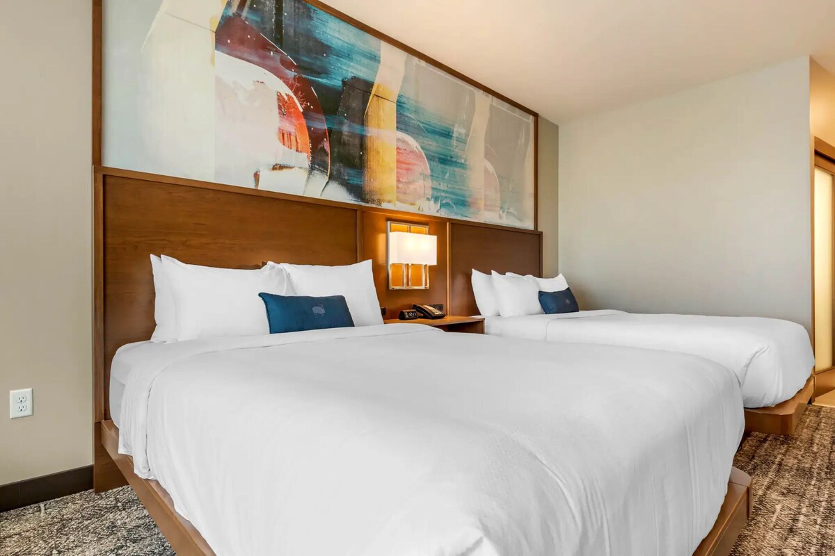 A hotel room with two beds, a large abstract painting, a bedside lamp, and a carpeted floor.