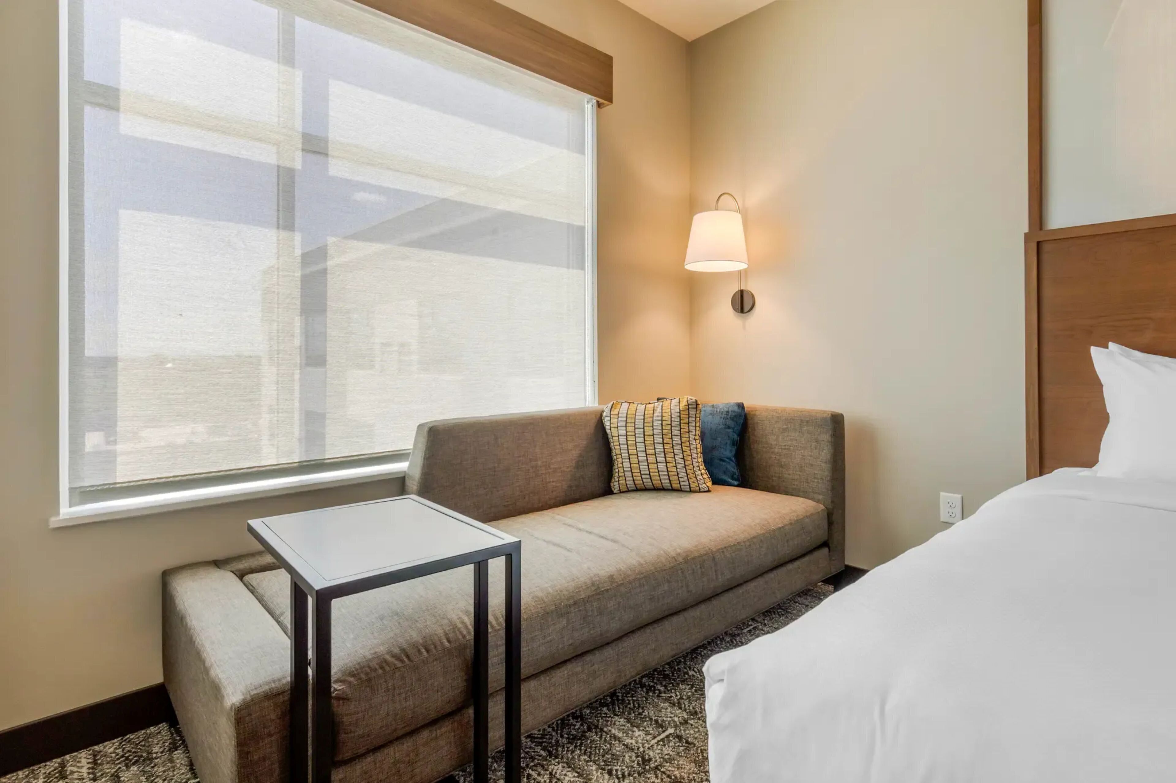 A hotel room with a sofa, a small table, a bed, a wall-mounted lamp, and a large window.