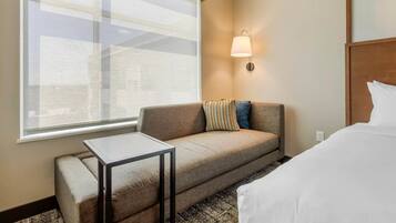 A hotel room with a sofa, a small table, a bed, a wall-mounted lamp, and a large window.