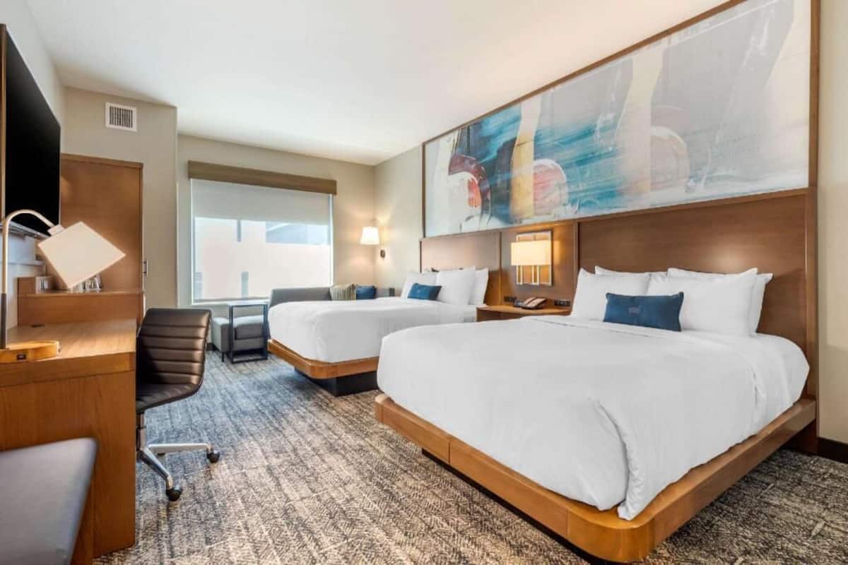 A hotel room with two beds, a desk, a chair, a TV, and a large abstract painting on the wall.