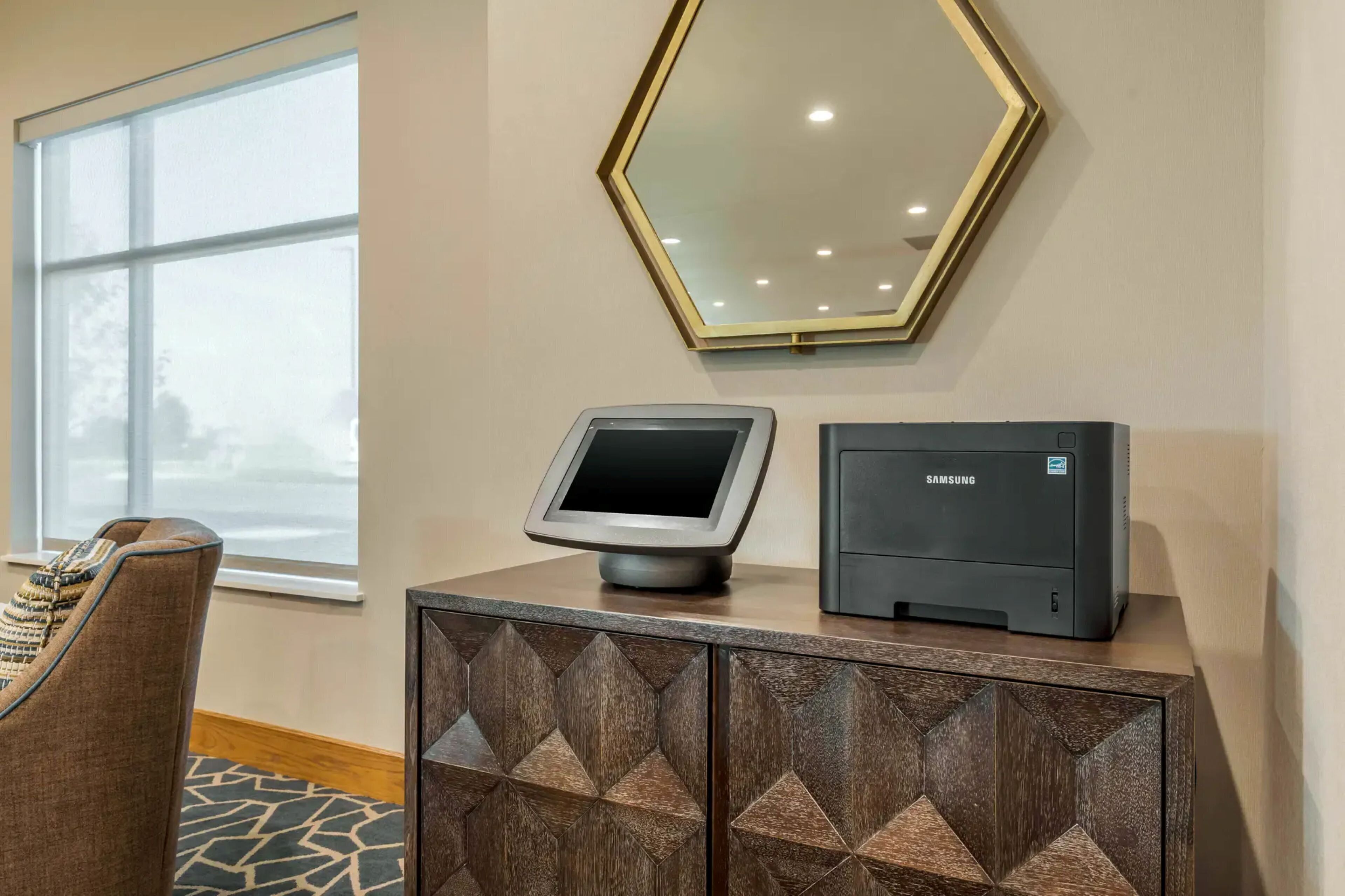 Una habitación con un amplio ventanal, un espejo en la pared, una impresora Samsung y un mueble con motivos.
