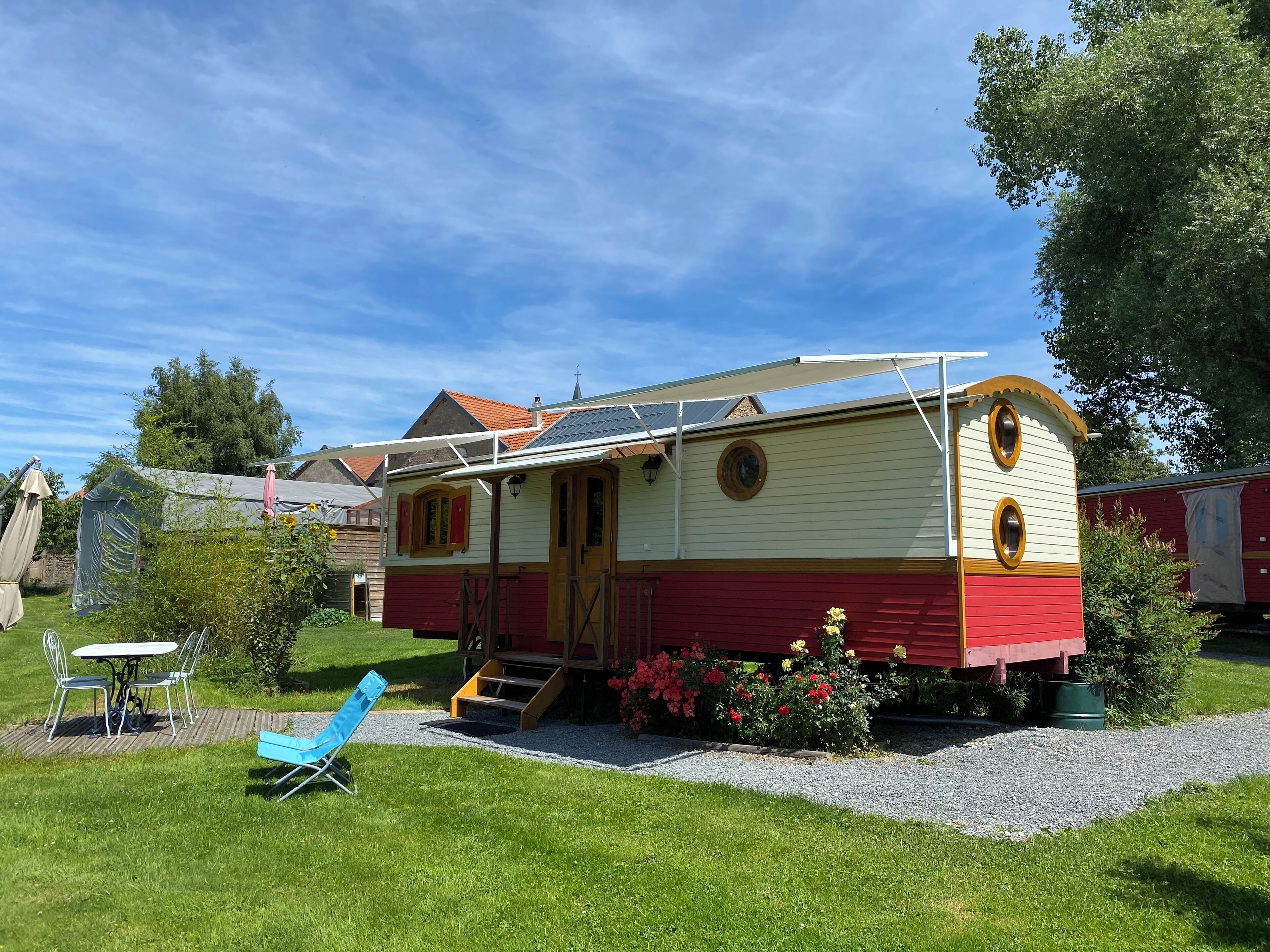Une petite maison à un étage, à l’extérieur rouge et crème, avec un porche à l’avant et un petit jardin agrémenté de fleurs et d’une chaise bleue.