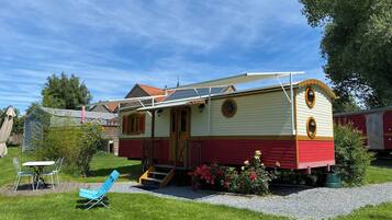 Une petite maison à un étage, à l’extérieur rouge et crème, avec un porche à l’avant et un petit jardin agrémenté de fleurs et d’une chaise bleue.