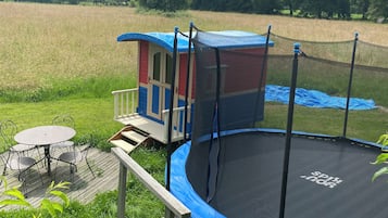 Un trampolí amb xarxa de seguretat, una caseta infantil i un pont de fusta amb taula i cadires.