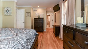 Une chambre à coucher avec une couvre-lit à motifs, une commode, un téléviseur fixé au mur et une porte donnant sur une autre pièce.