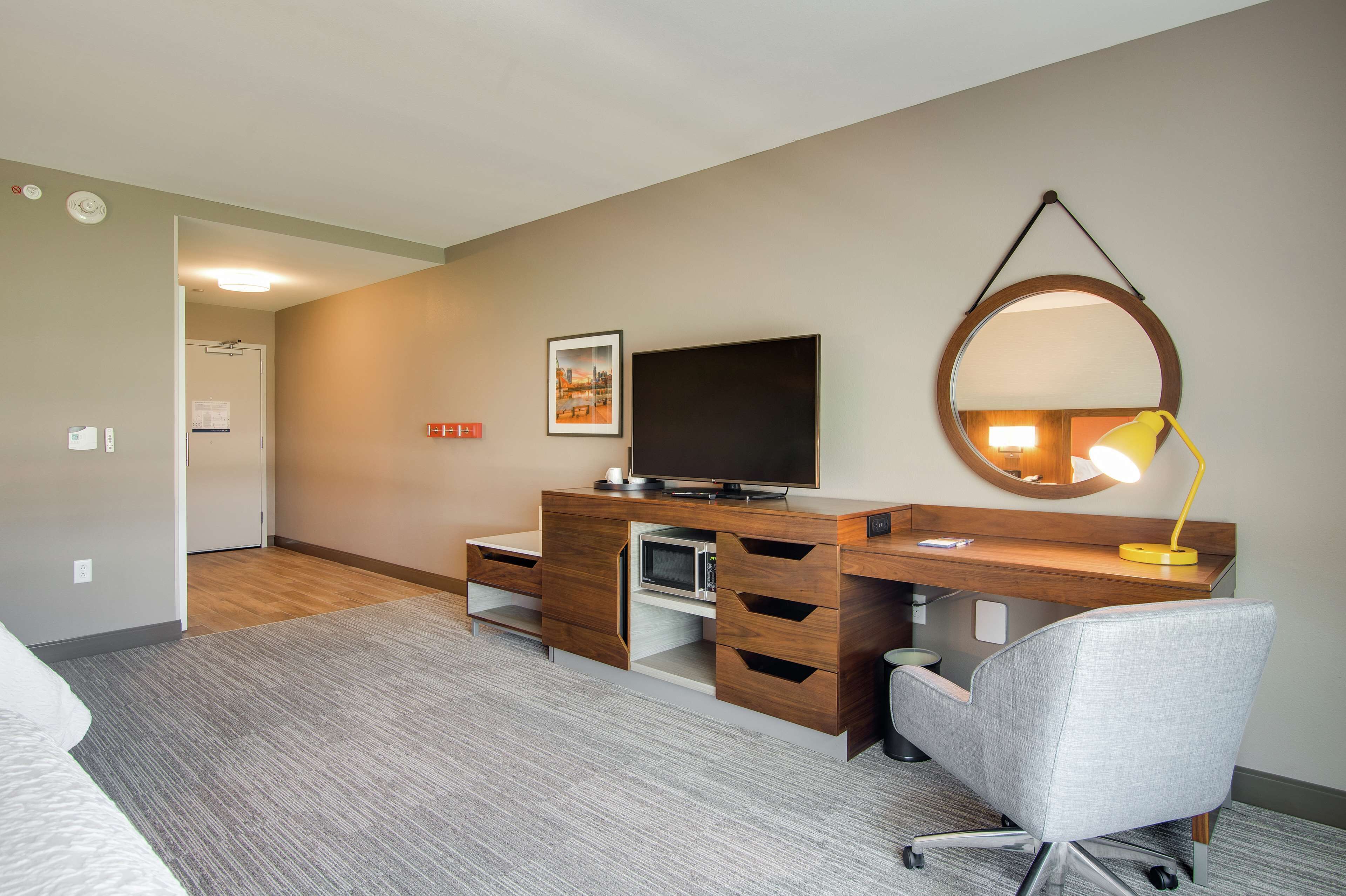 A modern hotel room with a wooden desk, a grey office chair, a flat-screen TV, a round mirror, and a yellow desk lamp.