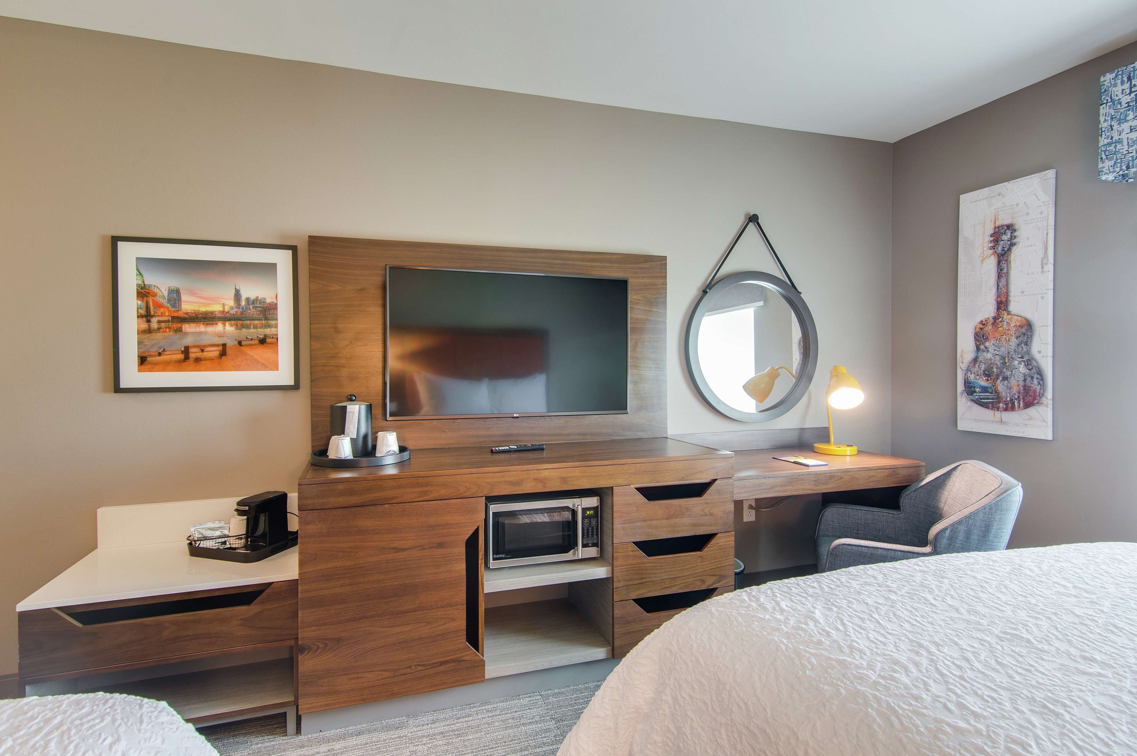 Habitación de hotel con escritorio de madera, televisor de pantalla plana, microondas, cafetera, lámpara y cama.