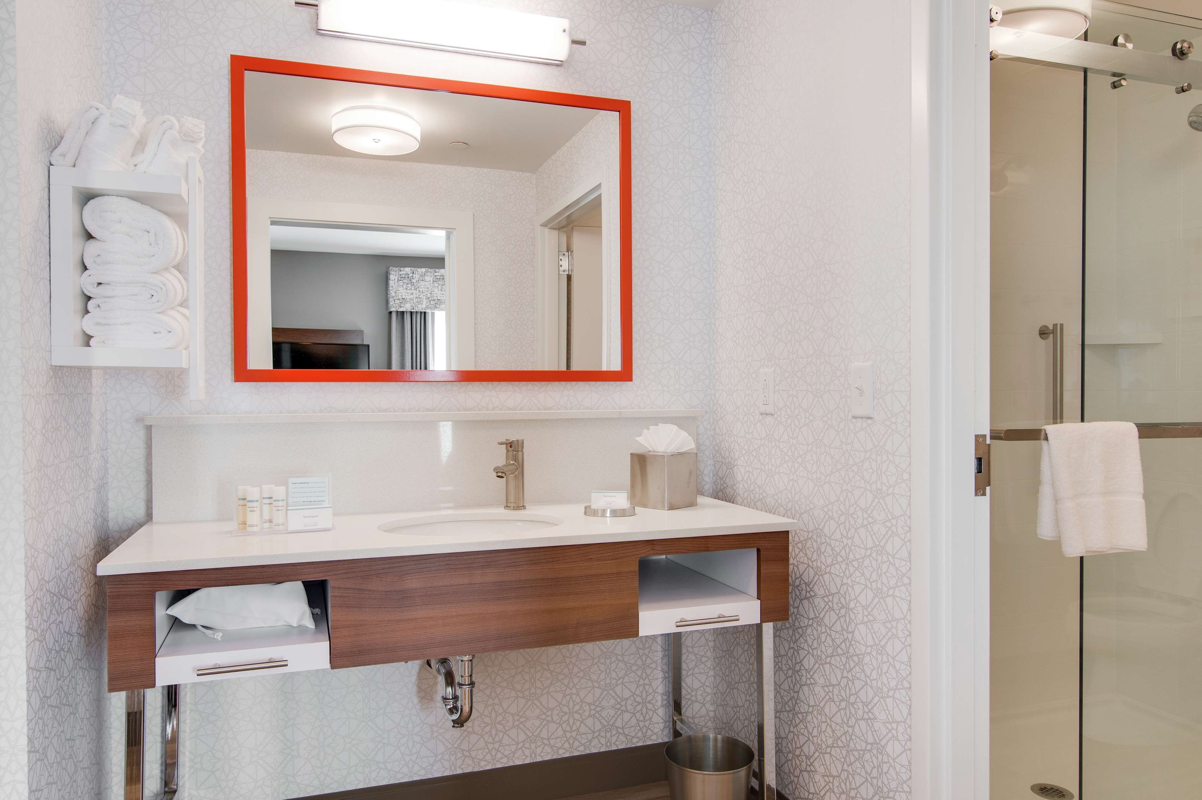 Un baño moderno con un lavabo blanco, un espejo con marco naranja, un perchero y una cabina de ducha con mampara de cristal.