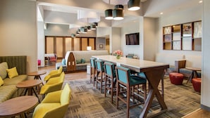 A modern hotel lobby with a long reception desk, multiple seating areas including a sofa and chairs, and a large wall-mounted TV.