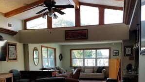 A spacious living area with a high ceiling, a ceiling fan, and large windows allowing natural light.