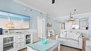 Un salón amplio con una gran televisión de pantalla plana, un sofá blanco y una zona de comedor con candelabro.