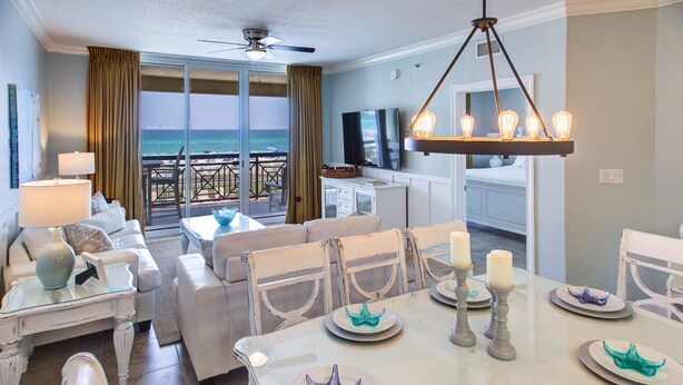 Un salón amplio con balcón con vistas al océano, amoblado con sofás blancos, una mesa de comedor con platos con forma de estrella de mar y una araña.