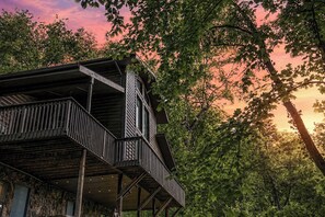 A wooden structure with a balcony, surrounded by trees and a sunset sky.