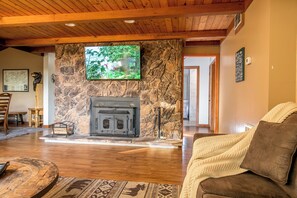 A cozy living room with a stone fireplace, wooden ceiling, and a television mounted on the wall.